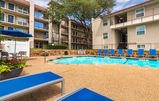 A pool area with lounge chairs, a patio table and chairs with a blue umbrella and stainless steel BBQ in front of apartment buildings at Princeton Court Apartments in the Vickery Midtown neighborhood of Dallas, TX.