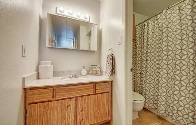 A tidy bathroom here at High Plains with a wooden vanity, light countertop, and a mirrored medicine cabinet with overhead lighting, featuring a bathtub-shower combo with a patterned curtain and a clean, functional layout.