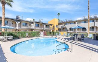 A sunny outdoor area featuring a sparkling blue swimming pool surrounded by lounge chairs and umbrellas, with palm trees in the background. The setting includes several buildings with balconies, and vibrant landscaping with flowers, creating a relaxing atmosphere.