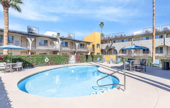 A sunny outdoor area featuring a sparkling blue swimming pool surrounded by lounge chairs and umbrellas, with palm trees in the background. The setting includes several buildings with balconies, and vibrant landscaping with flowers, creating a relaxing atmosphere.
