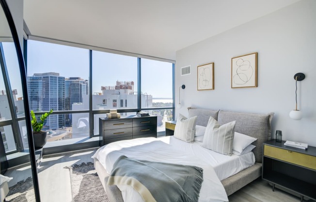 Bright and airy city-view bedroom with a comfortable bed adorned with white and grey pillows, flanked by minimalist nightstands and abstract wall art.