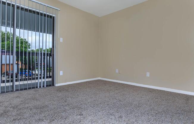 Empty room with beige walls and carpeted floor. There is a sliding glass door with vertical blinds, leading to a small balcony or outdoor area. Natural light is coming in through the door, illuminating the space. No furniture or decorations are visible.