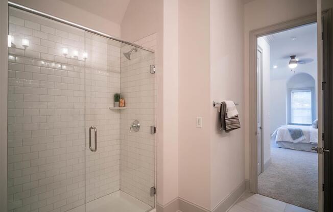 A glass-enclosed shower with subway tiles leads to a well-lit bedroom visible through an open door.