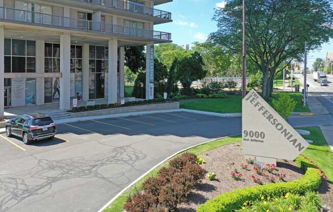 Outdoor image of the Jefferson. The parking lot is empty with the Jefferson sign next to the street. There are green bushes and blue skies.