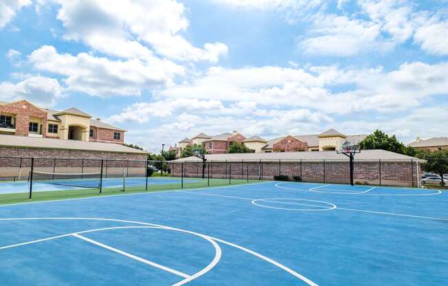 A basketball court with a blue surface and white lines is surrounded by a brick wall and houses.