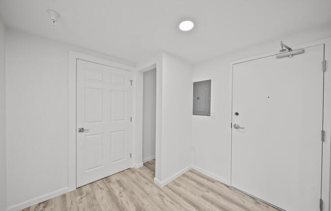 A modern, minimalist interior hallway with white walls and a light wooden floor. The space features a closed door on the left, a small closet or utility area in the center, and an entrance door on the right. A ceiling light illuminates the area, creating a bright and inviting atmosphere.