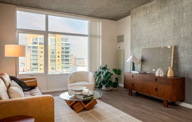 A living room with a brown sofa, a white chair, a wooden coffee table, and a large window with a view of a building outside.