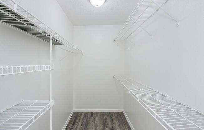 A spacious, empty closet featuring white wire shelving on both sides and a light fixture on the ceiling. The walls are painted white, and the floor has a wood-like design, creating a clean and bright storage area.
