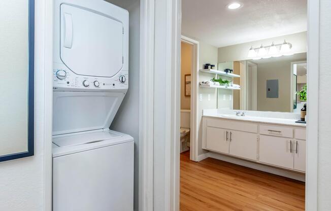 A compact laundry area featuring a stacked washer and dryer in a hallway. The space includes a bathroom visible through an open door, and a bright vanity with a mirror and shelves above. The flooring is wooden, and the walls are light-colored, creating a modern, clean aesthetic.