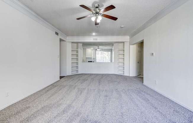 Spacious living area at Saxony at Chase Oaks Apartments in Dallas, TX, showcasing plush carpeted flooring, a ceiling fan with light, built-in shelving on either side of a mirrored accent wall, and a bright neutral color palette.