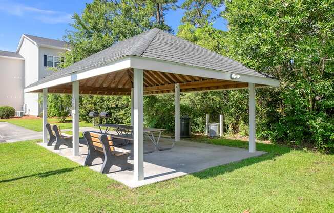 A pavilion with a roof and four benches is surrounded by greenery.