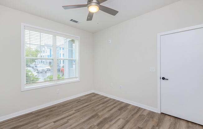 A vacant room featuring a ceiling fan, a window with blinds letting in natural light, and light-colored walls. The flooring is wooden, and there's a modern white door visible. Outside the window, residential buildings can be seen.