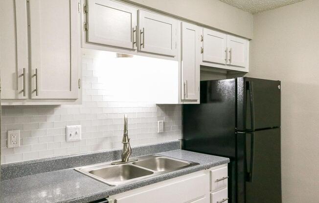 A modern kitchen featuring white cabinets, a granite countertop, and a black refrigerator. The kitchen includes a double sink with a sleek faucet and white subway tile backsplash, illuminated by overhead lighting. The overall design is clean and functional, suitable for everyday cooking.