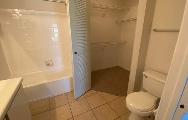 Bathroom with tile floors, toilet, shower tub combo with tile back splash, towel bar, white cabinets and view of closet with three mounted racks