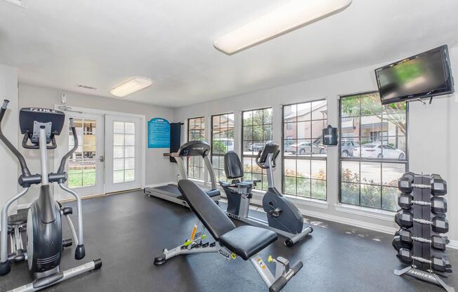 Bright gym interior featuring various exercise equipment, including treadmills, stationary bikes, and weight benches. Large windows provide natural light and a view of the outside, while a television is mounted on the wall. Weights are neatly stacked on the floor.