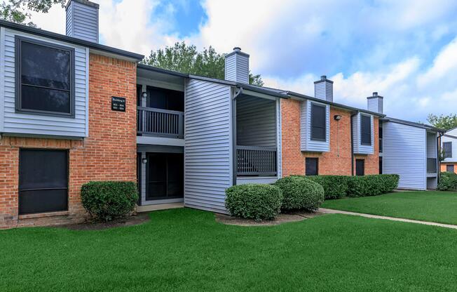 Two-story apartment building with a mix of brick and gray siding. Each unit has a balcony or patio, and there are several chimney-like structures on the roof. The grounds are well-maintained with lush green grass and neatly trimmed shrubs. The sky is partly cloudy.
