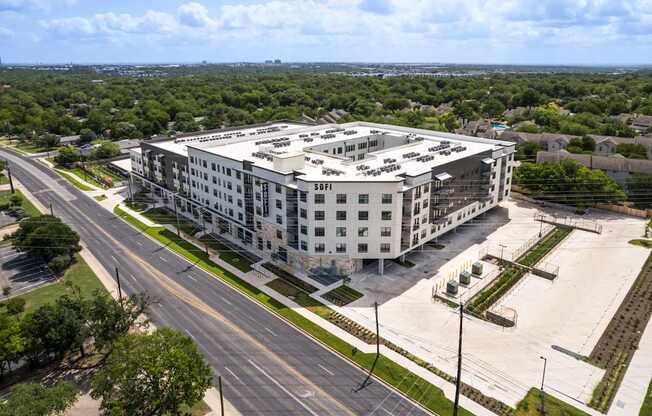 A large white building with a flat roof is surrounded by a parking lot and a road.