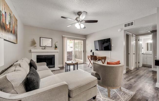 A cozy living room featuring a light gray sectional sofa, a modern chair, and a small table. There's a fireplace with decorative items, a flat-screen TV mounted on the wall, and large windows letting in natural light. A stylish area rug adds warmth to the space, which has neutral-colored walls and wood flooring.