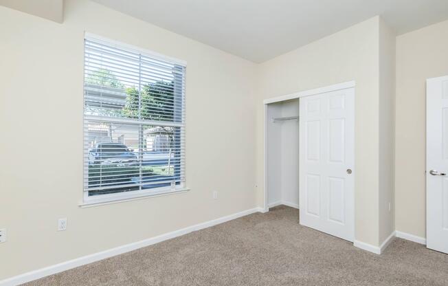 A well-lit room featuring a large window with blinds, beige walls, and carpeted flooring. On one side, there is a closet with sliding doors, and on the opposite side, a wall panel with white doors. The overall atmosphere is neat and inviting.
