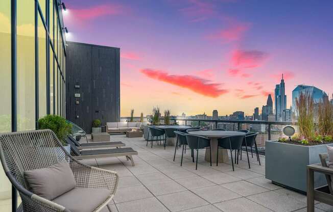 A rooftop patio with a table and chairs overlooking a city skyline at sunset.