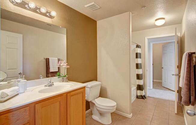 A warm and inviting bathroom here at Dobson Towne Center featuring a spacious vanity with wood cabinetry, a large framed mirror with bright overhead lighting, and a sleek white countertop with integrated sink. Neutral tile flooring leads to a full tub and shower combination, while soft accent walls and plush towels create a comfortable, well-appointed space.