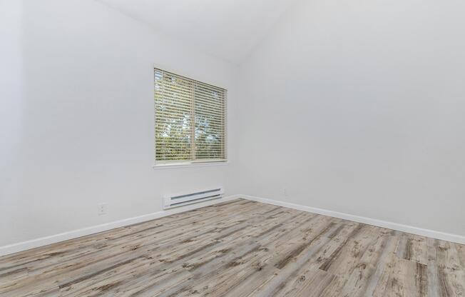 A bright, empty room with white walls and a high ceiling. There is a large window with blinds, allowing natural light to enter. The floor is covered in light-colored wooden laminate, creating a clean and spacious atmosphere. A heating vent is visible on the wall beneath the window.