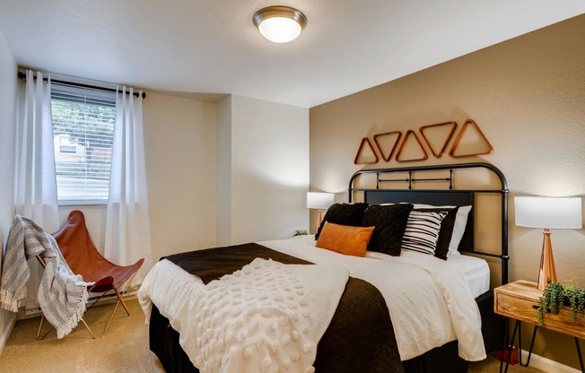 One Bedroom Apartments in Happy Valley, OR - Sunnyside Village - a Bedroom with Plush Carpeting, a Bed, Two Bedside Tables with Lamps, a Ceiling Lamp, and a Window with Binds and Curtains