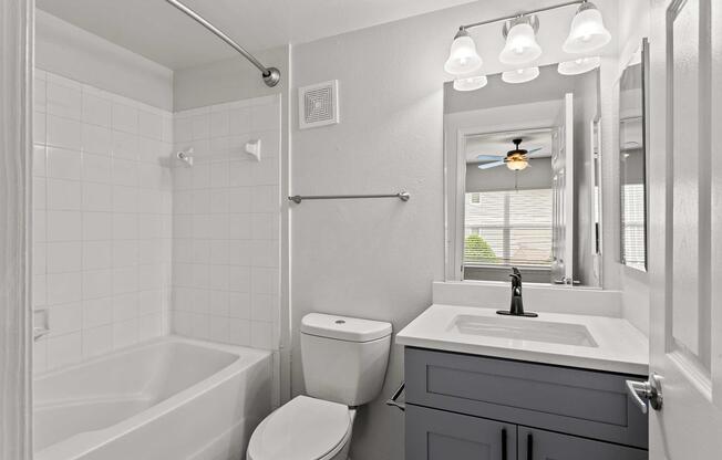 A clean and modern bathroom featuring a shower and bathtub combo, a white toilet, and a stylish gray vanity with a sink. The space is well-lit by overhead lights and has a mirror reflecting a window with blinds, enhancing the bright and airy feel of the room.