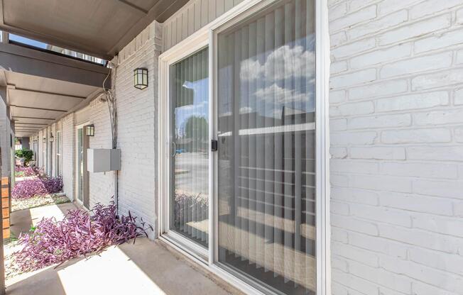 A view of a patio area featuring a sliding glass door with vertical blinds, surrounded by light gray brick walls. There is a small planter with purple foliage in front, and the scene is well-lit with blue skies and clouds visible through the glass.