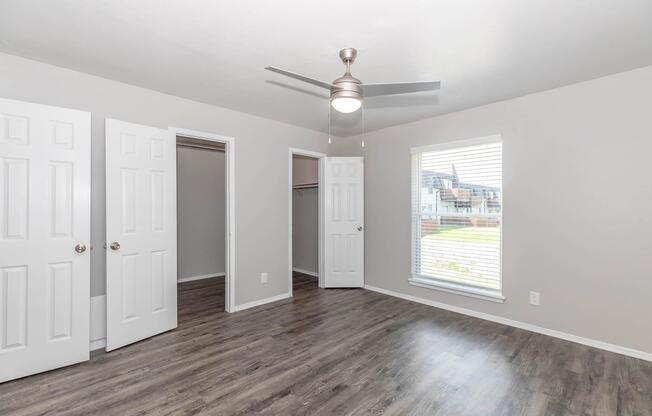 A spacious, well-lit bedroom featuring light gray walls, a ceiling fan, and laminate flooring. Two white doors lead to a closet and an adjoining room. A large window with blinds allows natural light to enter, and a neat, empty space gives a sense of openness.