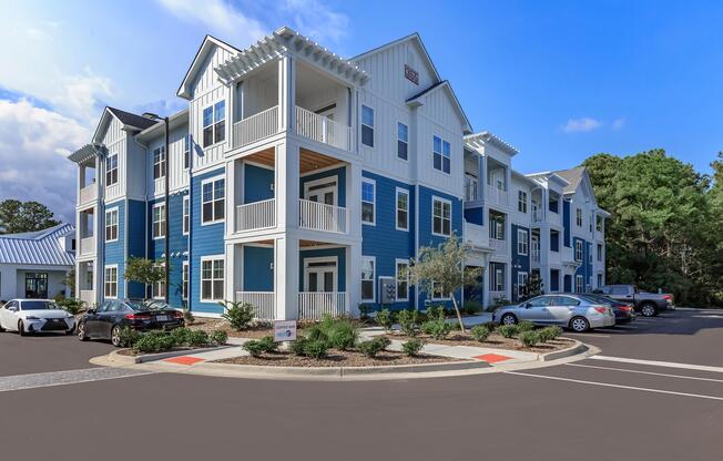 A modern, multi-story apartment building with a blue and white exterior. The building features balconies and landscaping with small trees and shrubs. There is a parking lot in front with several cars parked. The sky is partly cloudy, creating a bright and inviting atmosphere.