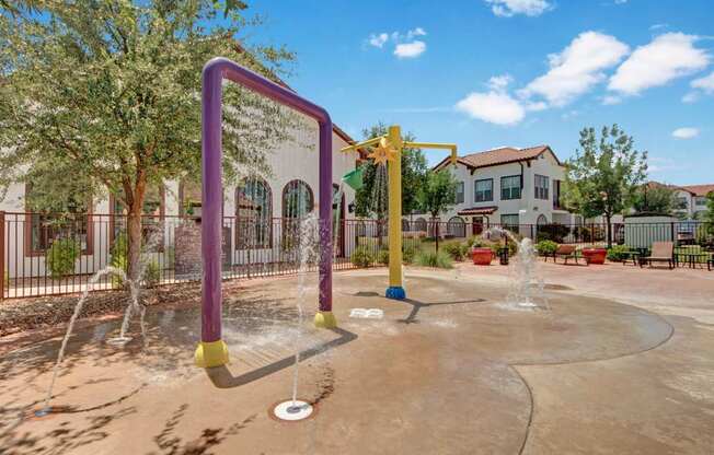 A colorful outdoor splash pad here at Andalucia Villas with playful water features, ground fountains, and shaded seating nearby, set against landscaped grounds and Spanish-style buildings for a fun, family-friendly atmosphere.