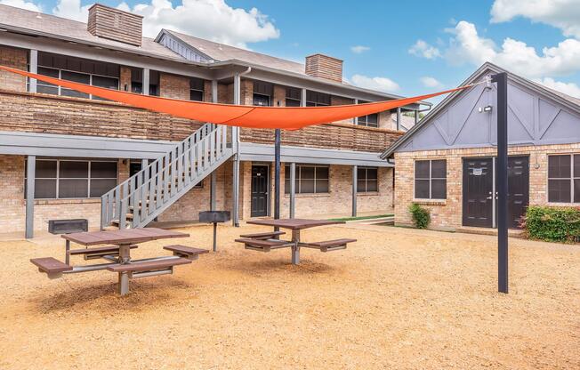 An outdoor area featuring several picnic tables shaded by an orange canopy. In the background, a two-story building with a wooden facade and multiple doors is visible. The ground is covered in gravel, and a staircase leads to the upper level of the building. Blue skies with fluffy clouds can be seen above.