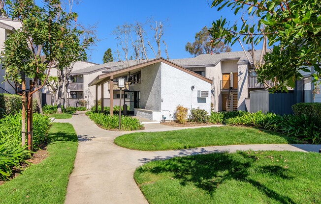 A white house with a grey roof is surrounded by greenery and a concrete pathway.