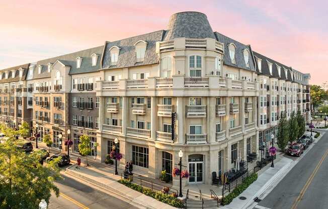 A large, multi-story building with a curved roof and balconies is situated on a street corner with a sidewalk and street lamps.