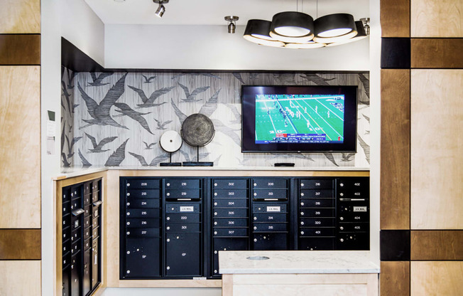 Interior view of a modern apartment complex mail room featuring rows of individual mailboxes neatly arranged along the wall. In the center, there is a flat-screen TV displaying important community announcements and updates.