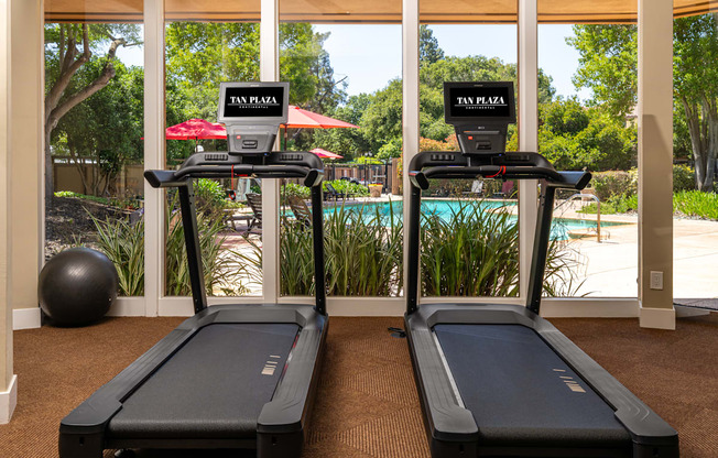 Two treadmills are placed in a room with a view of the pool and trees outside.