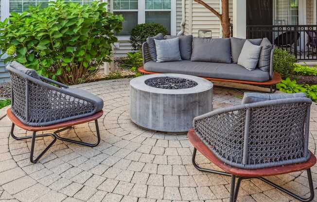 A grey couch with pillows sits on a patio with a fire pit in the middle.