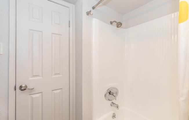 A clean, well-lit bathroom featuring a white shower stall with a chrome faucet and controls. There is a showerhead mounted on the wall and a white curtain partially visible. A closed, white door is seen next to the shower area, suggesting a tidy and modern design.