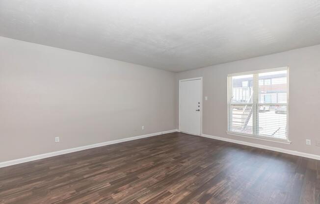 A bright, spacious room with light-colored walls and new wooden flooring. There is a large window allowing natural light to enter, and a door leading outside. The space is empty, offering a blank canvas for interior decoration.