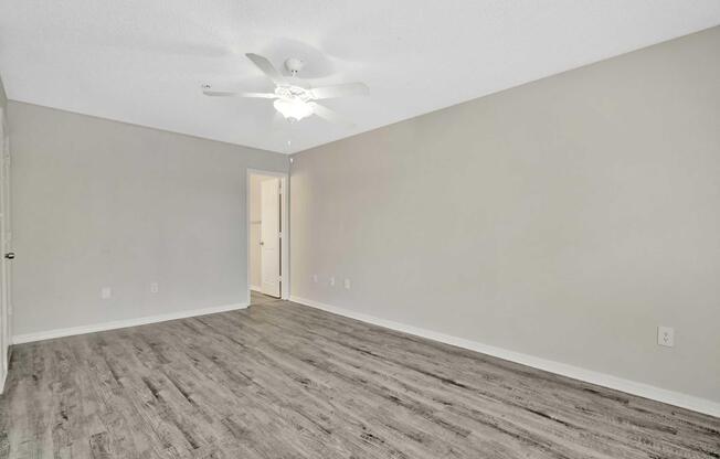 An empty room with light-colored walls and a ceiling fan. The floor is covered with light wood laminate. A doorway is visible on the right side, leading to another space. The room is well-lit, creating a spacious and airy atmosphere.