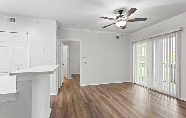 Interior view of a modern apartment living space featuring a ceiling fan, light-colored walls, and laminate flooring. On one side, there is a sliding glass door with vertical blinds, and on the other, a white partition leads to a doorway. The area is well-lit, creating a clean and open atmosphere.