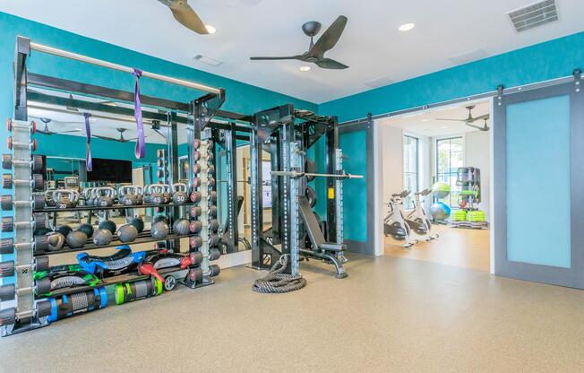 A well-lit gym area featuring a variety of fitness equipment. On one side, there are free weights and resistance bands neatly arranged. A multi-use strength training machine stands prominently. The other side shows an entrance to another fitness area with exercise bikes and yoga mats. The walls are painted in a vibrant teal color.