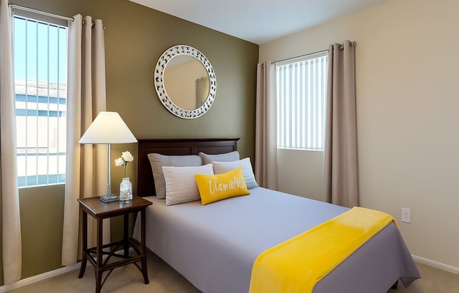 A warm and inviting bedroom here at Pala Mesa featuring soft carpeting, a rich wood headboard, and neutral bedding accented with a vibrant yellow throw and decorative pillow. A bedside table with lamp adds cozy lighting, while two tall windows with vertical blinds and curtains fill the space with natural light.