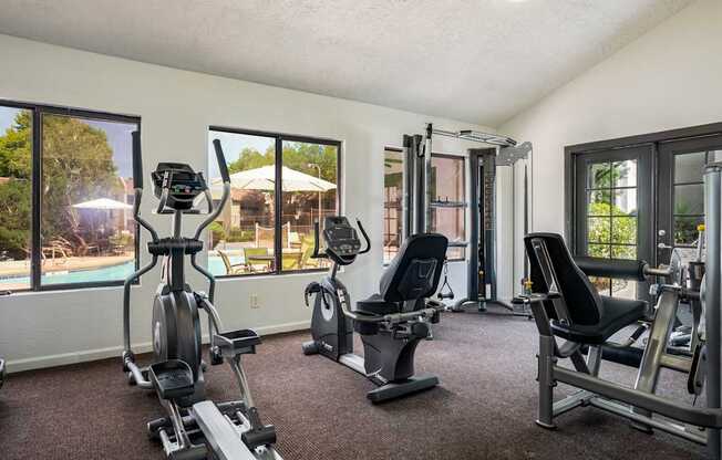 A well-equipped gym with a view of a pool and trees outside the windows.