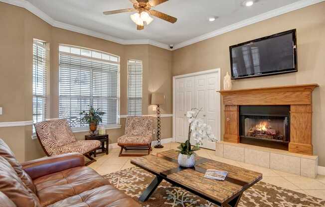 A warm, inviting living room here at Mission Green featuring a cozy fireplace with a wooden mantel, mounted television, plush seating, accent chairs by large windows with blinds, a patterned area rug, coffee table décor, ceiling fan, and soft neutral finishes that create a comfortable space for relaxing and gathering.