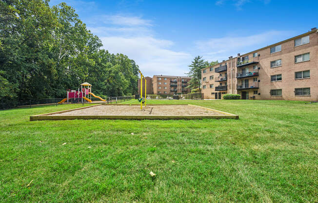 A playground with a slide and a yellow structure is in the foreground of a grassy area with apartment buildings in the background.