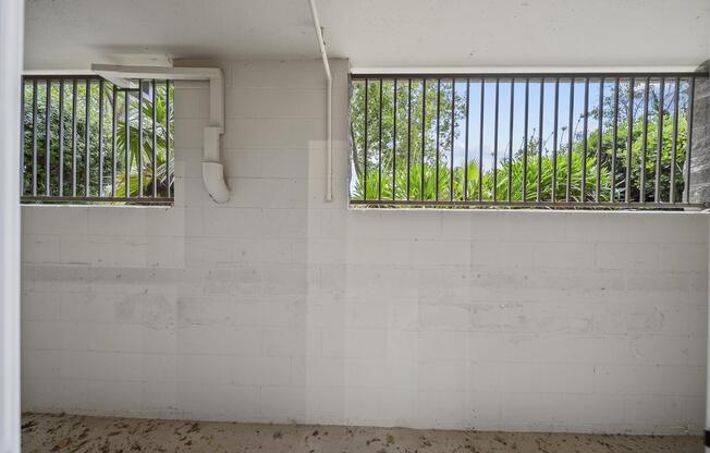 An empty interior space with two barred windows allowing light in. The walls are painted white with visible concrete blocks. Outside the windows, green foliage can be seen. The floor is bare, indicating a lack of furnishings or decoration.