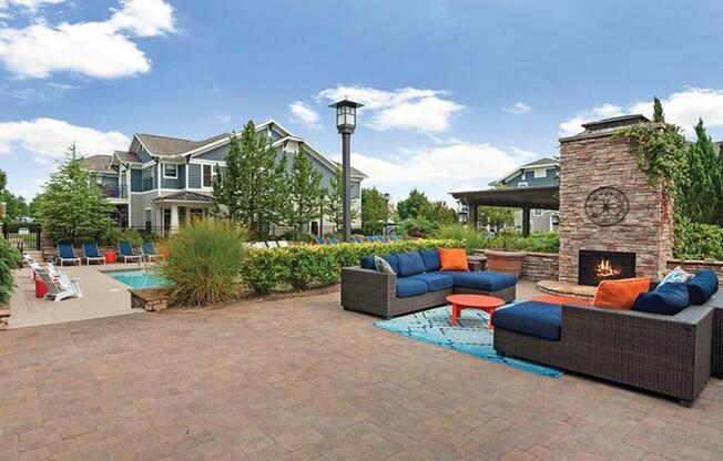 outdoor living room with gas fire place on a stone patio located next to pool area and lush landscaping