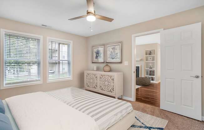 A virtually staged bedroom with the living room located in the background. The bedroom features gray walls with white trim, carpet throughout, two single windows with blinds, a three blade ceiling fan, and a door that opens to the living room.